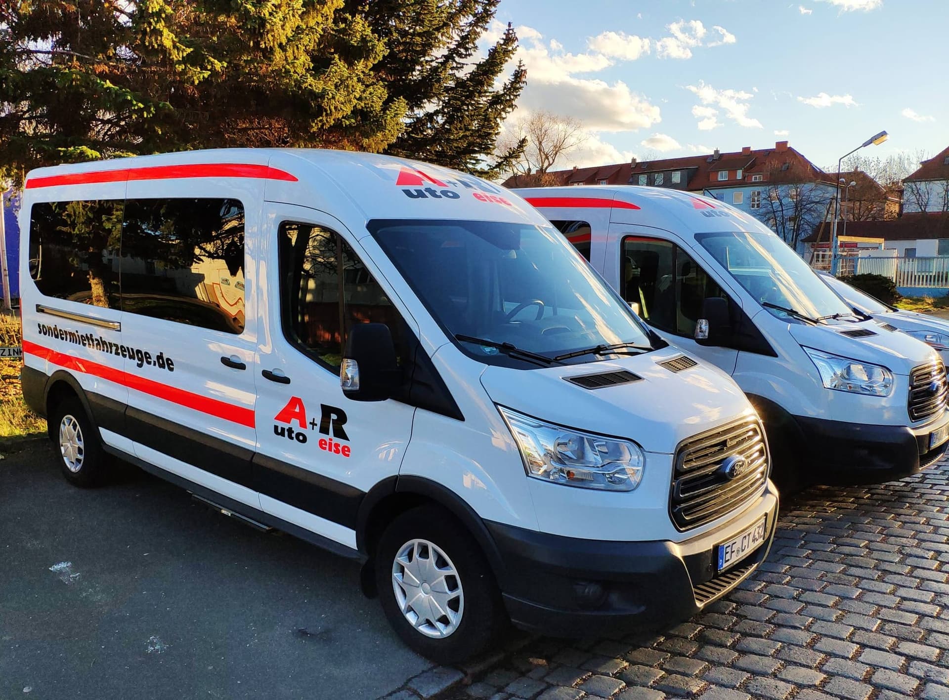Zwei weiße Ford Transit Kleinbusse mit roten Streifen und A+R-Beschriftung auf einem gepflasterten Hof